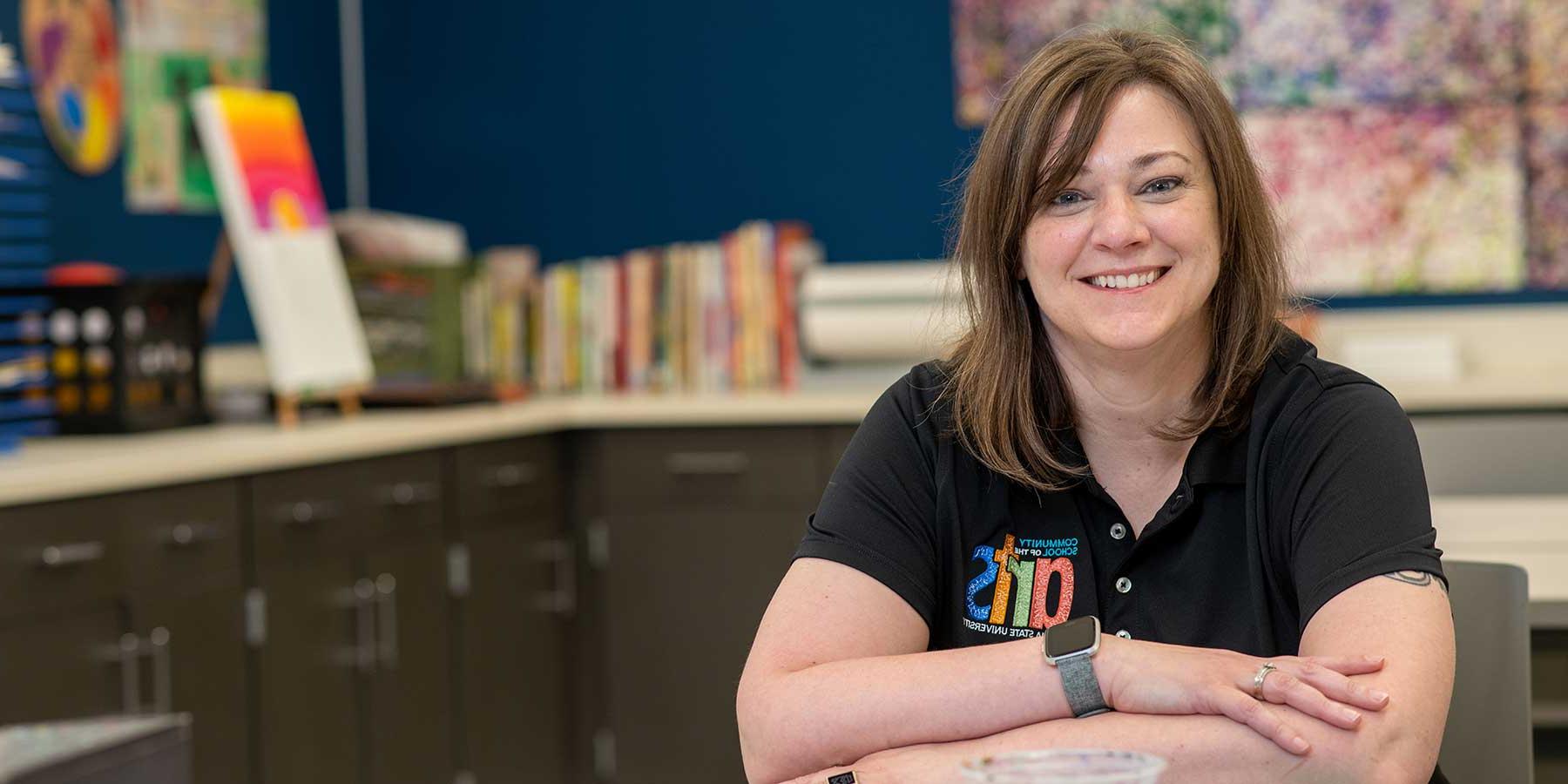 A white woman with shoulder-length brown hair sits, smiling at the camera. She wears a black T-shirt with a colorful patch reading Community School of the Arts, Indiana State University. Cabinets and counters are visible behind her with art supplies and art projects